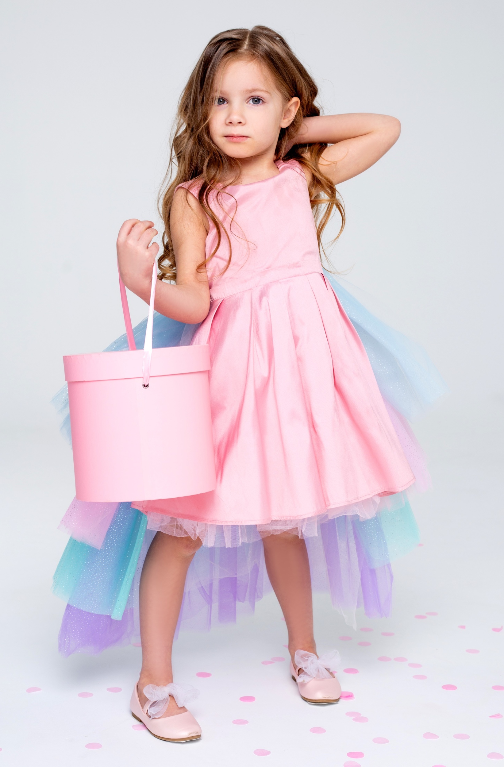 3 krasivaa-malen-kaa-devocka-v-sikarnom-rozovom-plat-e-poziruet-stoa-na-serom-fone-i-derzit-slapnuu-korobku-v.jpg